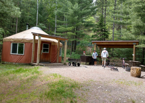A yurt in a forested area with two people near a picnic table and a bike, surrounded by trees.