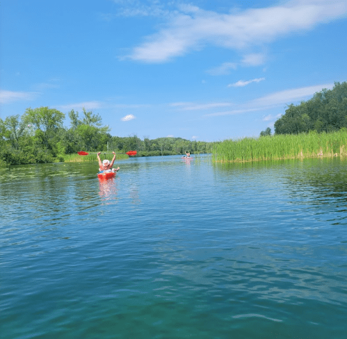 A person in a red kayak raises their arms in a serene, sunny river surrounded by greenery and blue skies.