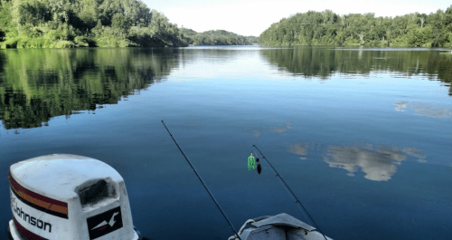 A calm lake scene from a boat, with fishing rods and lush greenery reflected in the water.