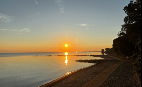 Sunrise over a calm lake, with a sandy shore and trees lining the water's edge, reflecting warm colors in the sky.