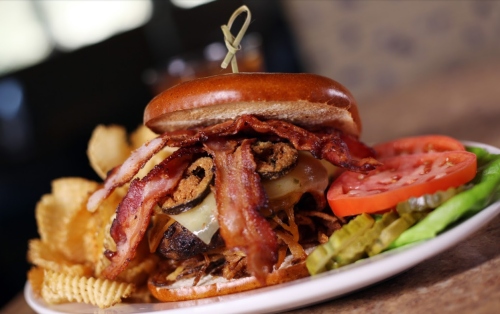 A juicy burger topped with bacon, cheese, and fried onions, served with chips, tomato, lettuce, and pickles on the side.