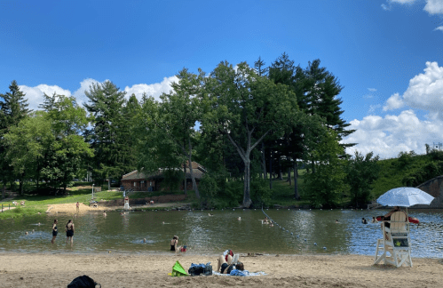 A sandy beach by a calm lake, with people swimming, lounging, and a lifeguard chair under a blue sky with clouds.