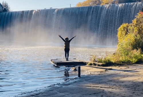 A person stands on a dock with arms raised, facing a waterfall surrounded by trees and mist.