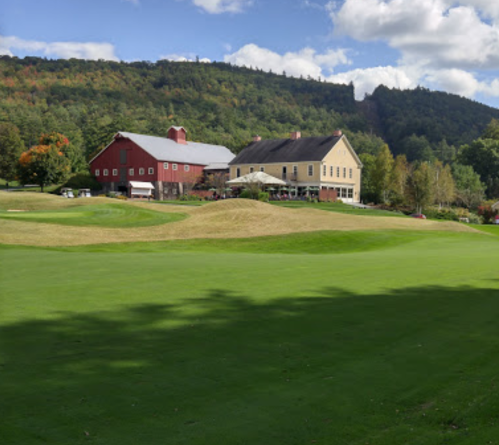 A scenic view of a golf course with a red barn and a large house against a backdrop of green hills and blue skies.