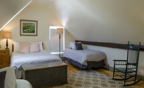 Cozy attic bedroom with two beds, a rocking chair, a lamp, and a window, featuring warm, inviting decor.
