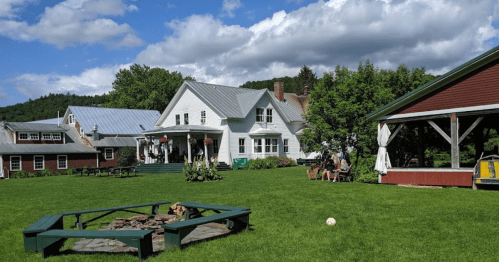 A scenic view of a green lawn with a fire pit, surrounded by rustic buildings and trees under a partly cloudy sky.