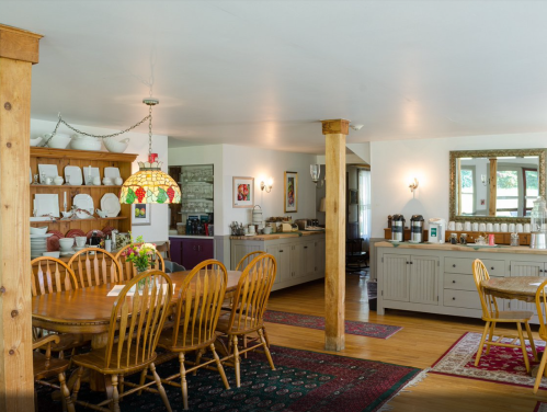 A cozy dining area with a large wooden table, chairs, and a colorful lamp, featuring shelves and a buffet in the background.