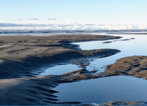 A serene landscape featuring a winding waterway through sandy terrain, with snow-capped mountains in the background.