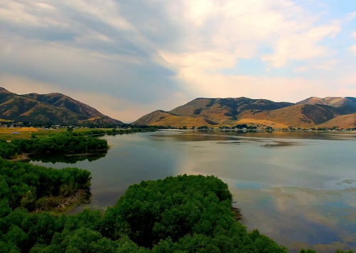 A serene landscape featuring a calm lake surrounded by green trees and mountains under a cloudy sky.