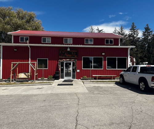 A red building with a sign reading "Sydney's," featuring large windows and a parking lot with a white truck.