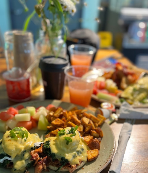 A plate of eggs Benedict with greens, potatoes, and fruit, with drinks and a floral centerpiece in the background.