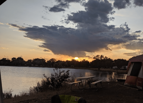 Sunset over a calm lake, with clouds in the sky and a picnic table near a camping tent in the foreground.