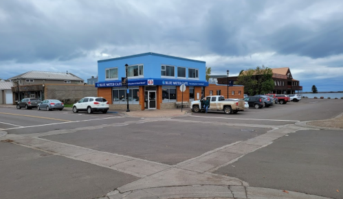 A blue building with a sign for "Blue Water Cafe" near a parking lot and cloudy sky. Cars are parked nearby.