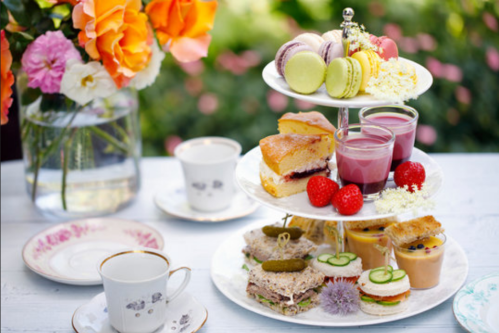 A tiered stand with colorful desserts, sandwiches, and drinks, alongside floral arrangements and elegant teacups.