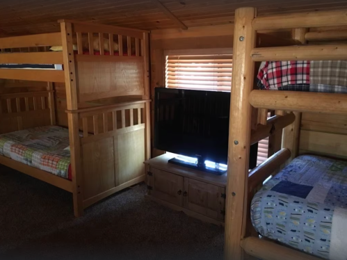 Cozy cabin bedroom with two sets of wooden bunk beds and a TV on a wooden stand, illuminated by natural light.