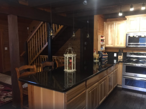 A rustic kitchen with wooden cabinets, a black countertop, and a staircase in the background. Lanterns hang from the ceiling.