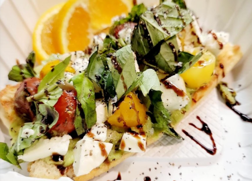 A colorful flatbread topped with fresh greens, tomatoes, cheese, and drizzled with balsamic glaze, served with orange slices.