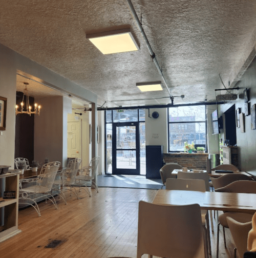 A cozy café interior with wooden floors, light fixtures, and seating areas, featuring large windows and a welcoming atmosphere.