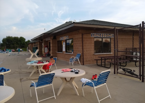 A poolside concession stand with tables and chairs, surrounded by lounge chairs and a fence. A child is sitting nearby.