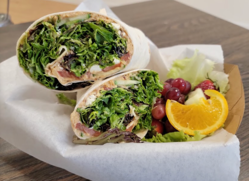 A fresh wrap filled with greens and sliced tomatoes, served with a side of mixed fruit and orange slices.