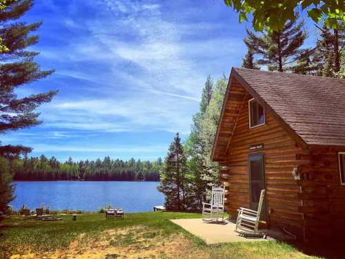 A cozy log cabin by a serene lake, surrounded by lush trees and a clear blue sky. Rocking chairs on the porch invite relaxation.