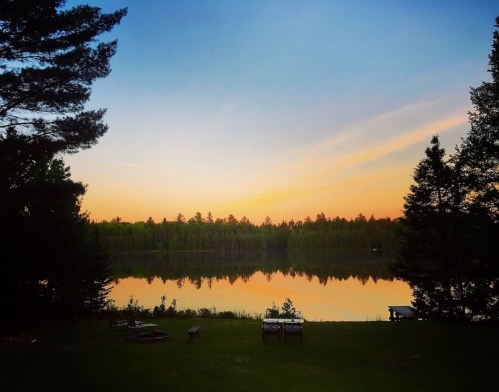 A serene lake at sunset, reflecting colorful skies and surrounded by lush trees. Cozy seating area in the foreground.