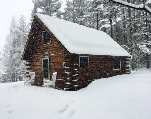 A cozy log cabin covered in snow, surrounded by tall trees, with rocking chairs on the porch. Snowflakes are falling.