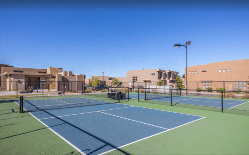 Two tennis courts with blue surfaces and surrounding buildings under a clear blue sky.