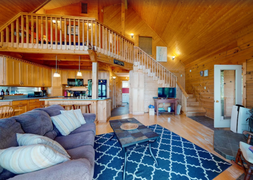Cozy wooden interior of a cabin with a living area, kitchen, and staircase, featuring a sofa and a patterned rug.