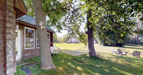 A peaceful outdoor scene featuring a hammock between two trees, with a cozy cabin and lawn chairs nearby.