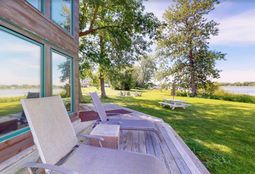 A serene lakeside view with lounge chairs on a wooden deck, surrounded by green grass and trees.