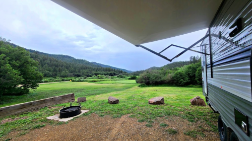 A scenic view from an RV, showing green fields and mountains under a cloudy sky, with a fire pit in the foreground.