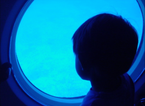 A child gazes out of a round window into a deep blue underwater scene.