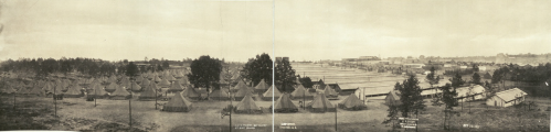 A panoramic view of a military camp with numerous tents and buildings, surrounded by trees and open fields.