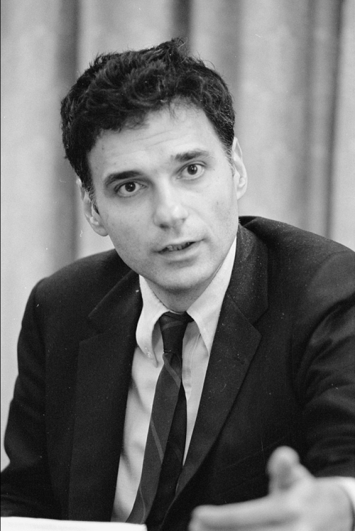 A man in a suit and tie gestures while speaking, with a serious expression and a blurred background.