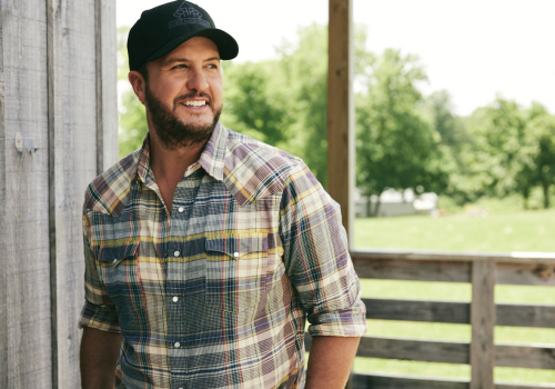 A man with a beard smiles while wearing a plaid shirt and cap, standing outdoors near a wooden structure.