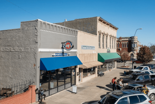 A street view of a small town with shops, including a grill, under a clear blue sky. Cars are parked along the sidewalk.