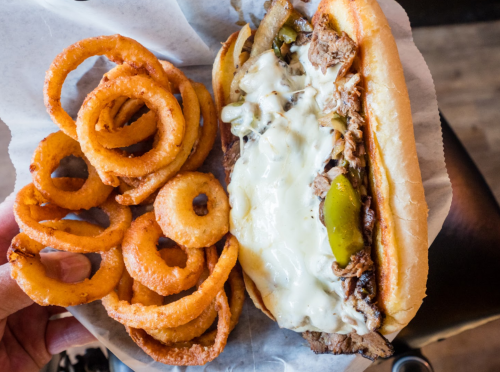 A hand holding a sandwich filled with beef, cheese, and peppers, alongside a serving of crispy curly fries.