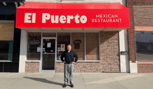 A man stands in front of "El Puerto Mexican Restaurant," featuring a bright red awning and an open sign.