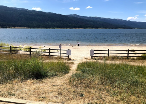 A sandy beach by a lake, framed by grass and wooden fences, with a person standing near the water under a blue sky.
