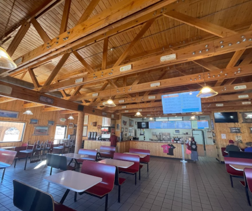 Interior of a rustic restaurant with wooden beams, tables, and a counter displaying a menu on a screen.