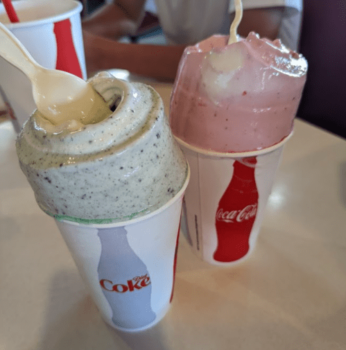 Two cups of thick milkshakes, one green with cookie bits and one pink, with spoons resting on top, on a table.