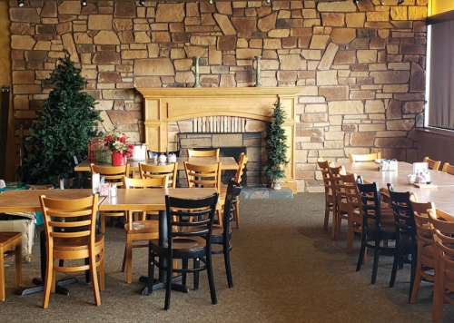 Cozy restaurant interior with wooden tables, chairs, and a stone fireplace, flanked by small evergreen trees.