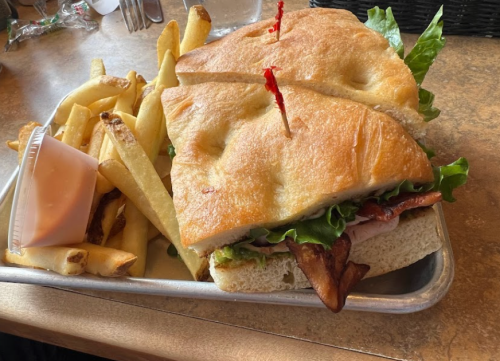 A sandwich with lettuce, bacon, and ham on a tray, accompanied by a side of French fries and a small container of sauce.