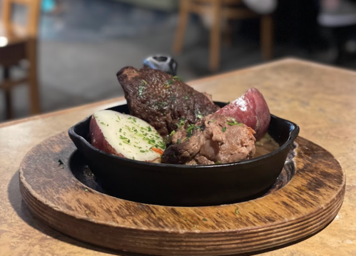 A sizzling skillet with beef, potatoes, and herbs, served on a wooden board in a cozy dining setting.