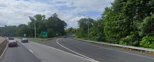 A highway exit with green signs, surrounded by trees and cloudy skies, with cars driving on the road.