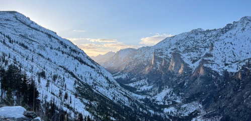 A snow-covered mountain valley at sunset, with rugged peaks and a clear sky in the background.
