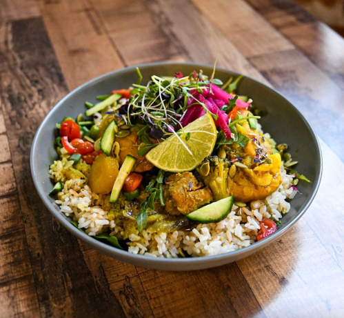 A bowl of colorful rice topped with vegetables, herbs, lime, and garnishes on a wooden table.