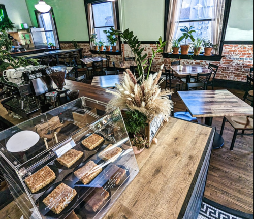 A cozy café interior with wooden tables, plants, and a display case of baked goods.