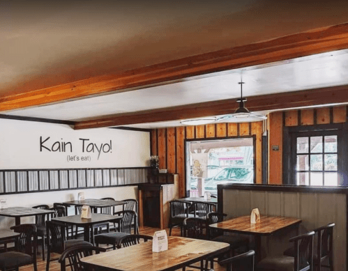 Cozy restaurant interior with wooden tables and chairs, featuring a "Kain Tayo!" sign on the wall.